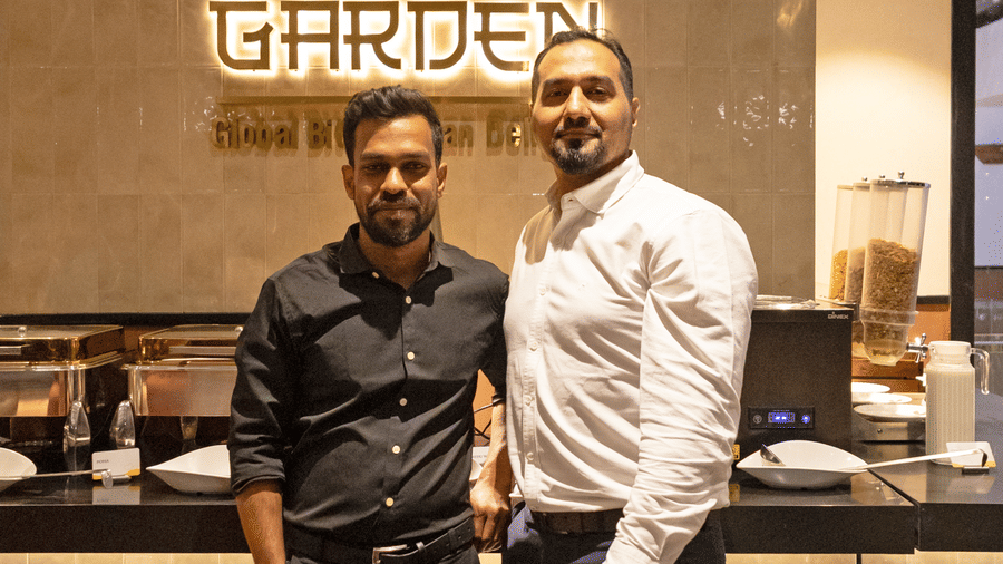  Two men stand side by side posing for a photograph inside Koi Garden at Hotel Bawa Gurgaon. The restaurant logo is mounted on the wall behind them above a counter structure.