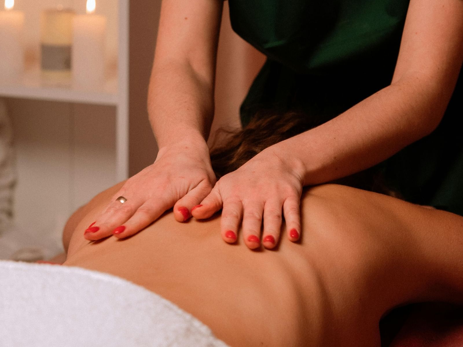 Close-up of a massage therapist in a green uniform with red nail polish giving a back massage to a client on a massage table with lit candles nearby.