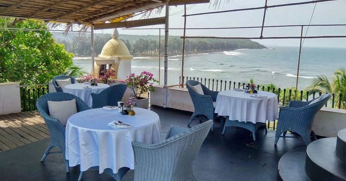 An outdoor dining area at Fort Tiracol, Goa, with tables set with white tablecloths and light blue chairs, under a shaded canopy. 