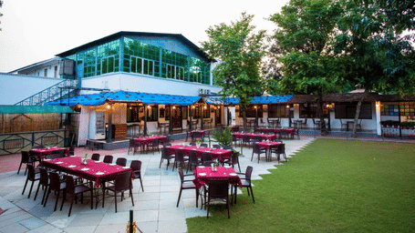 An outdoor dining setup featuring multiple red-clothed tables on a patio next to a green grass lawn and a building structure at Amantra Shilpi Resort, Udaipur.