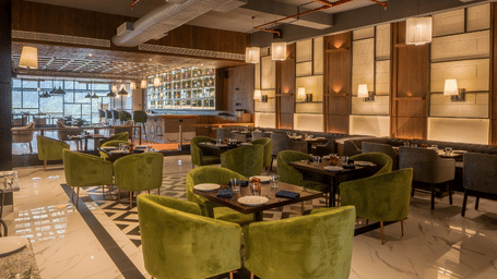 Restaurant with green velvet chairs, marble floor, exposed ducts and bar shelving in the background at Polo Hotel, Agartala.