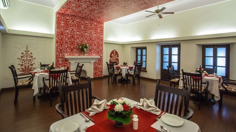 An indoor dining hall at Royal Heritage Haveli with high ceilings, several tables with white tablecloths, and a decorative red ceiling.