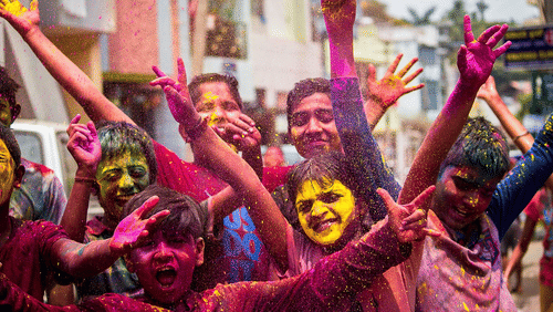 A group of people celebrating Holi, smiling and raising hands, with faces and clothes covered in coloured powder.