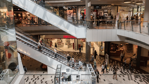 An interior of a shopping mall with multiple floors, escalators, people walking, and retail stores on each level.