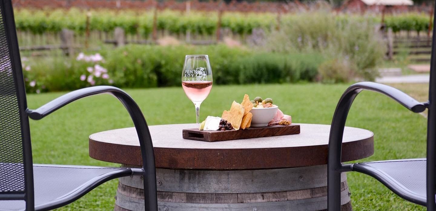 A glass of rosé wine and a charcuterie board sit on a barrel between two chairs