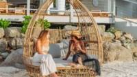 A beachside scene features a swing set on the sand with buildings and lush greenery in the background.