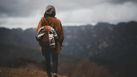 A lone hiker with a backpack walking along a mountain trail on a cloudy day, surrounded by misty hills and rugged natural scenery.