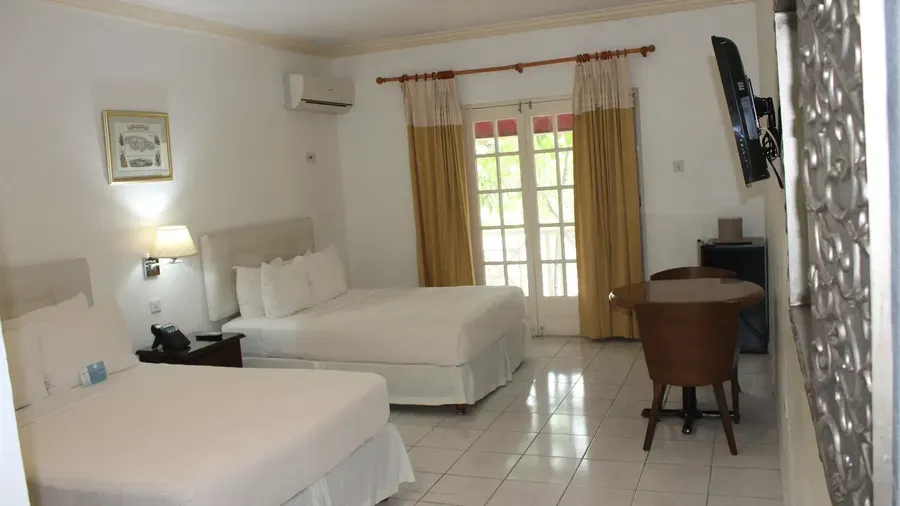 A clean hotel room includes a bed with white linens, a dark wooden dresser, and a window with light-colored curtains | Deluxe Double Room | Altamont Court, Jamaica