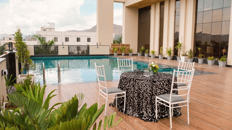 Several chairs placed with a table infront of a swimming pool at Pride Premier Alwar.
