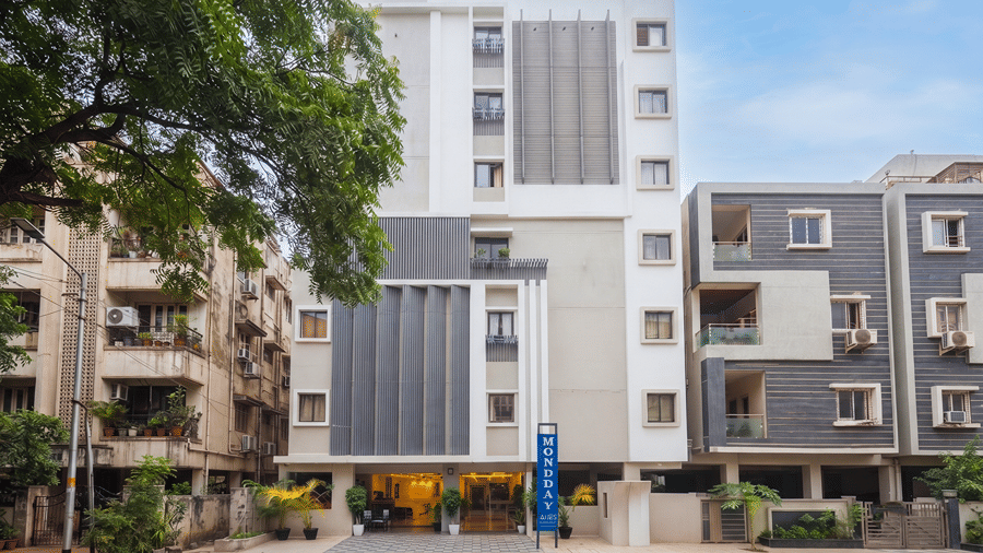 Front facade of Monday Hotels & Suites Madhapur, Hyderabad in daylight, with a blue vertical sign, lit entrance, potted palms, trees and neighbouring buildings.