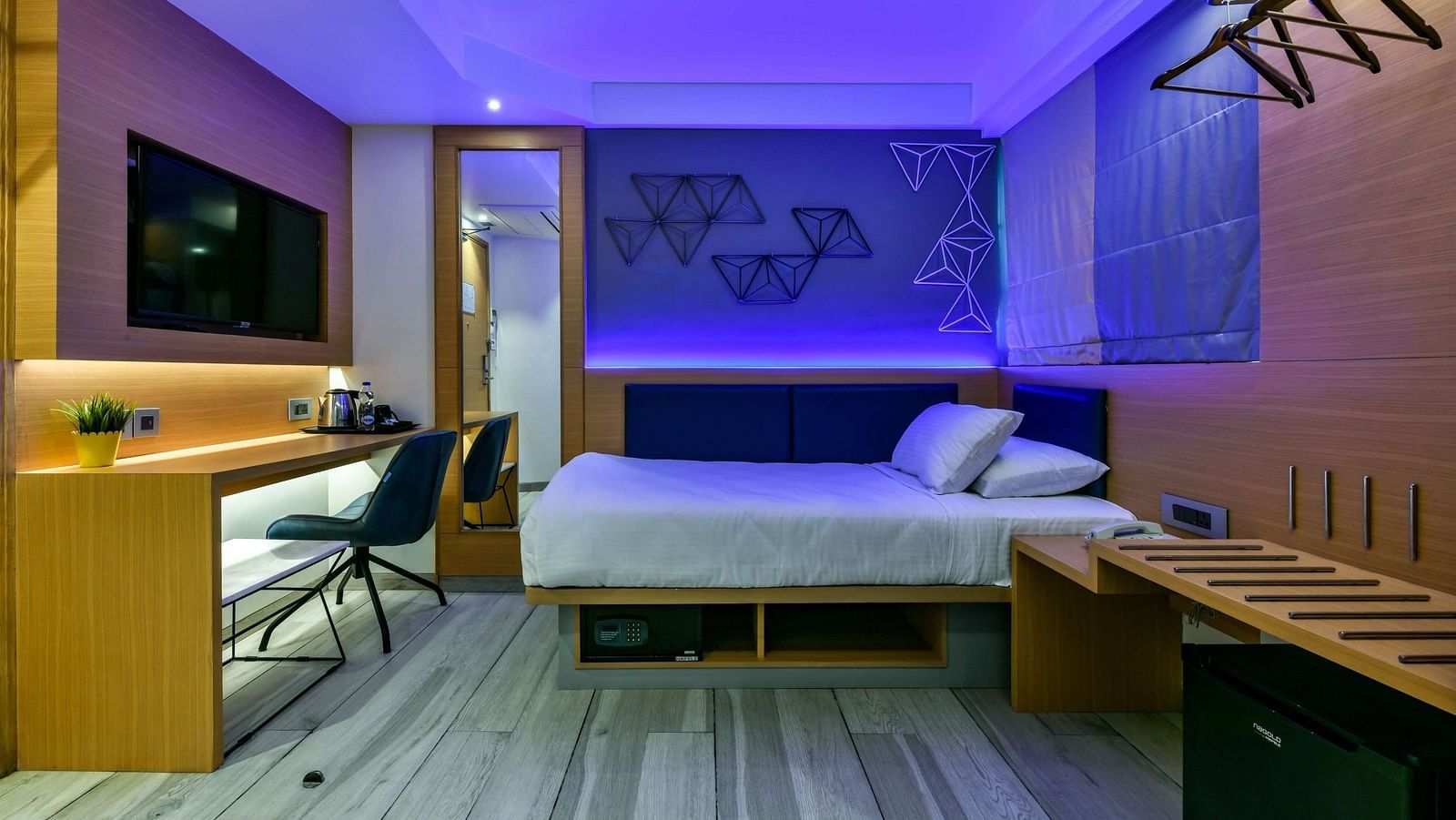 A spacious room featuring a bed, a wooden desk with a chair, a wall-mounted TV, and cool lighting at Indie Stays, BKC Kalina.