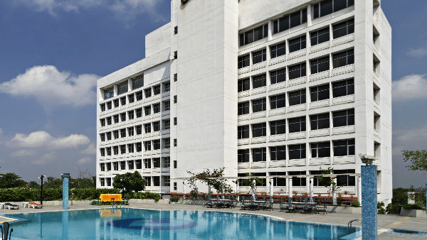 A large, modern white hotel building is shown behind a spacious blue swimming pool with lounge chairs on the patio - Clarks Avadh, Lucknow