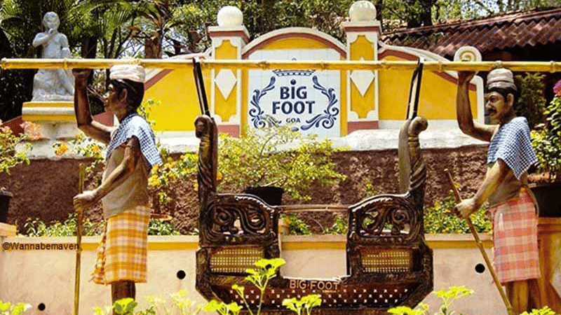 The entrance of Big Foot museum with a yellow sign, flanked by two statues wearing traditional clothes and hoisting a palanquin.