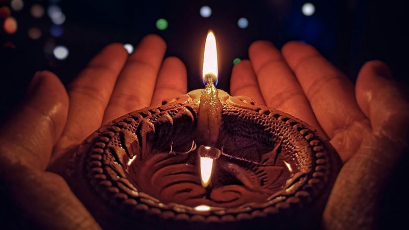 A person's hands cup a traditional clay lamp with a single, bright flame in the center. The background is a dark, blurry field of colourful, out-of-focus lights.