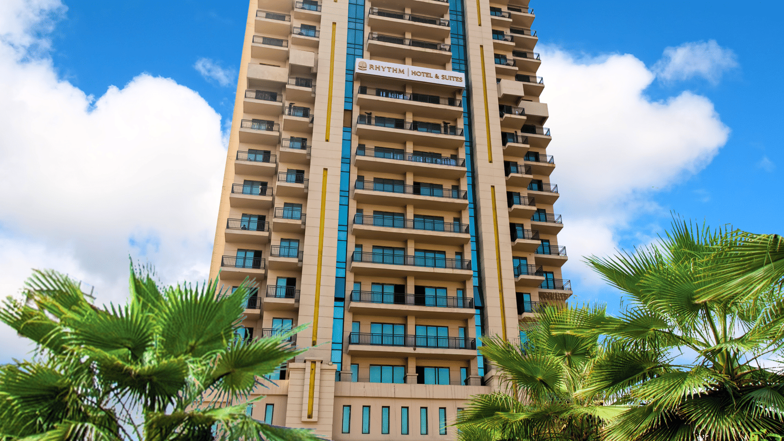 Front facade of Rhythm Gurugram surrounded by palm trees and landscaped greenery.