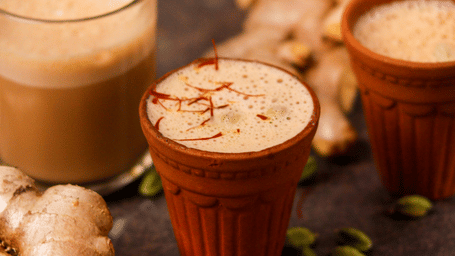Clay cups filled with tea placed on a dark surface with cardamom, ginger, and a grater scattered around.