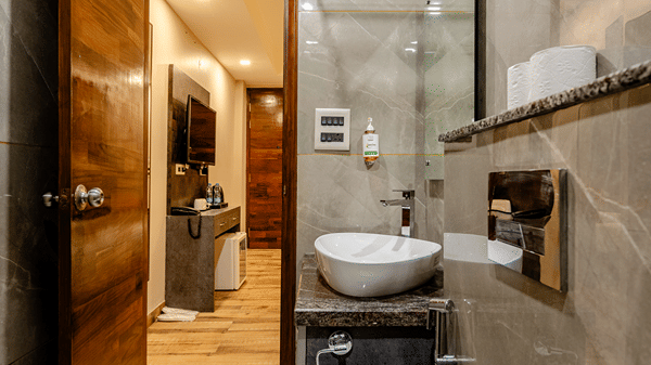 Sleek hotel ensuite bathroom with a glass shower screen, grey wall tiles, and an open wooden entry door.