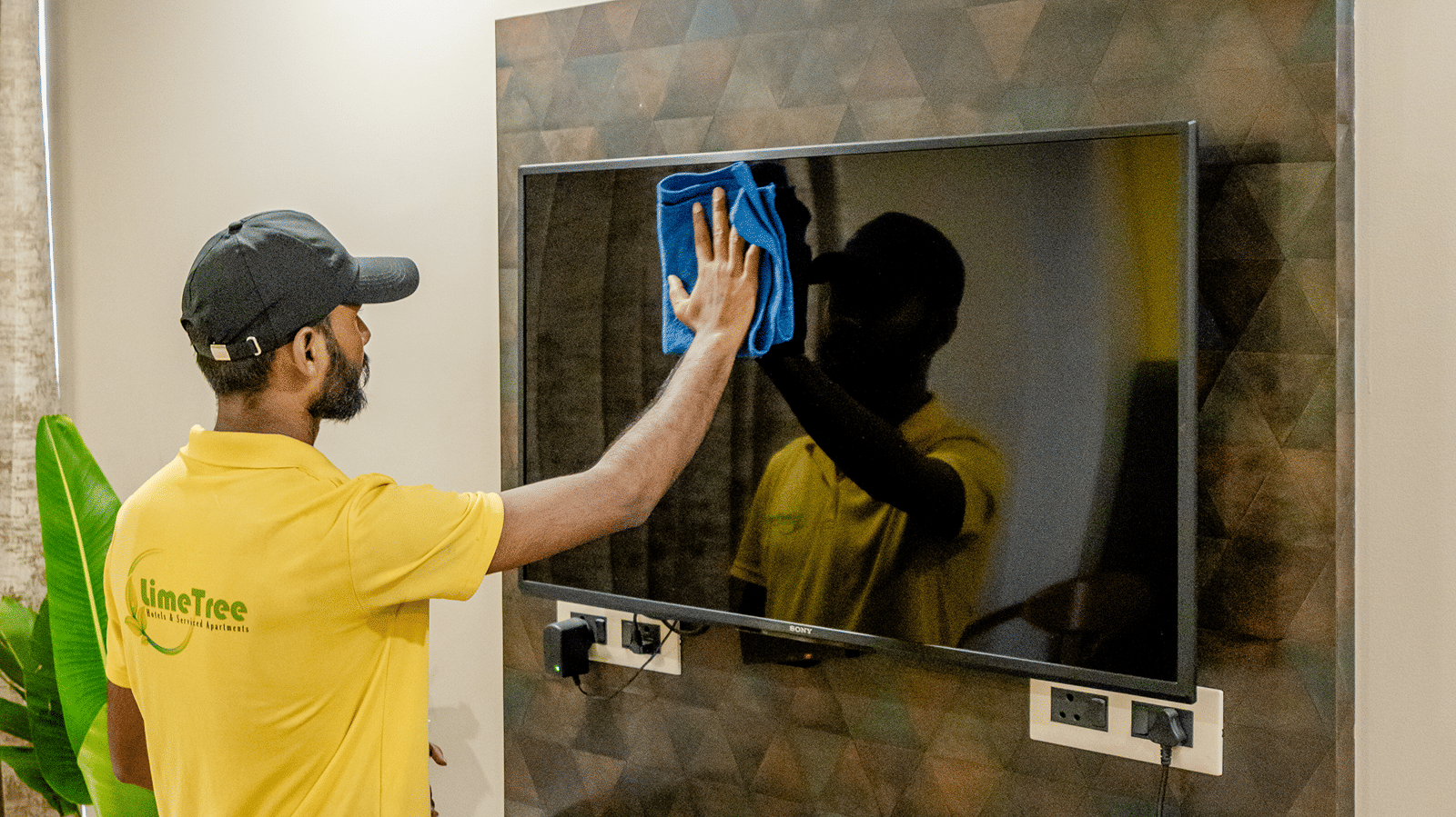 Hotel staff member in a cap and yellow shirt wiping a flat-screen TV on a dark textured accent wall.