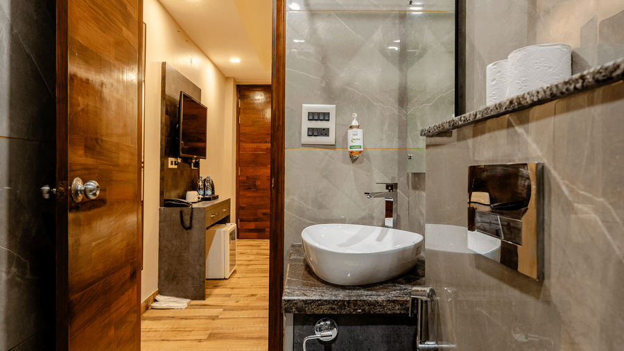 Sleek hotel ensuite bathroom with a glass shower screen, grey wall tiles, and an open wooden entry door.