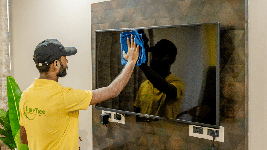 Close-up of a hotel staff member wearing a cap and yellow shirt wiping down a flat-screen television on a dark textured accent wall.