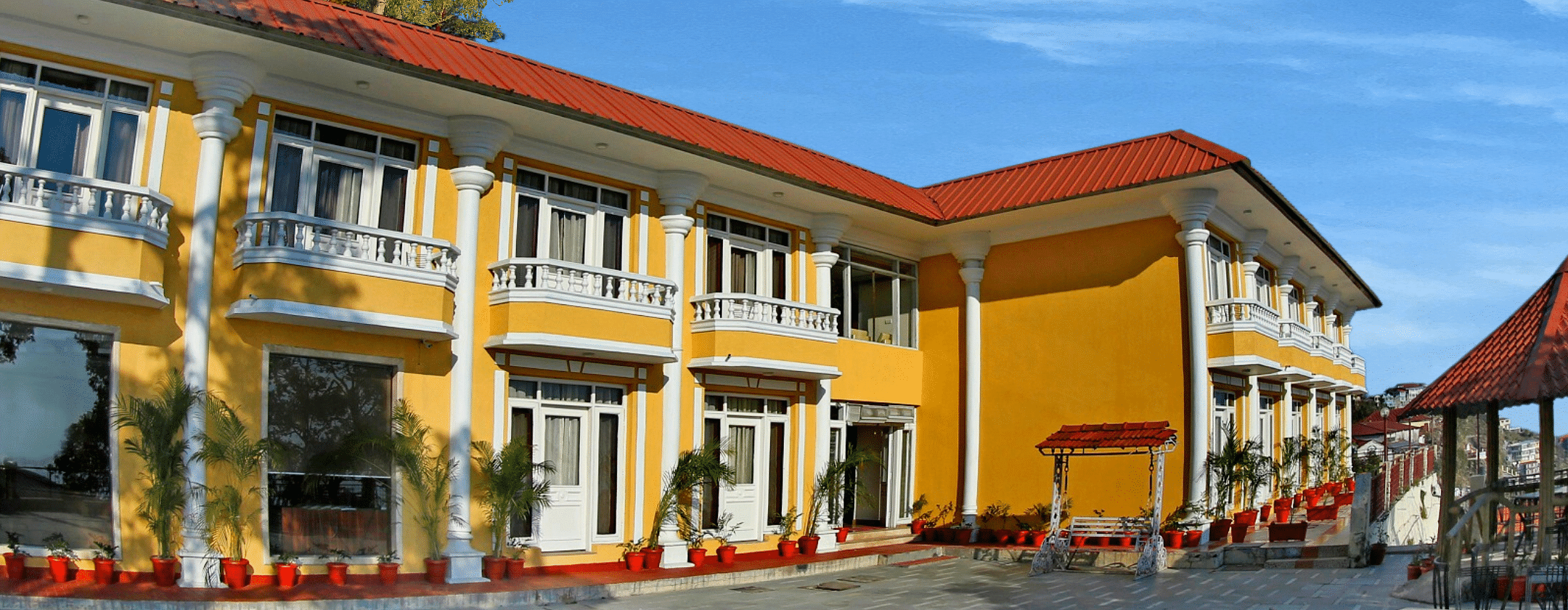 A corner exterior view of the  building with a roof and a balcony, surrounded by trees and a clear blue sky.