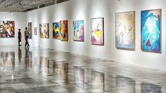 The image shows an art gallery with a long, white-walled hallway with colorful paintings are hanging on the walls