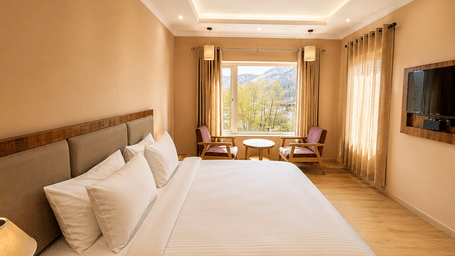 Hotel room with a bed dressed in white linen, a seating area with wooden chairs, a wall-mounted television and a large mountain-view window.