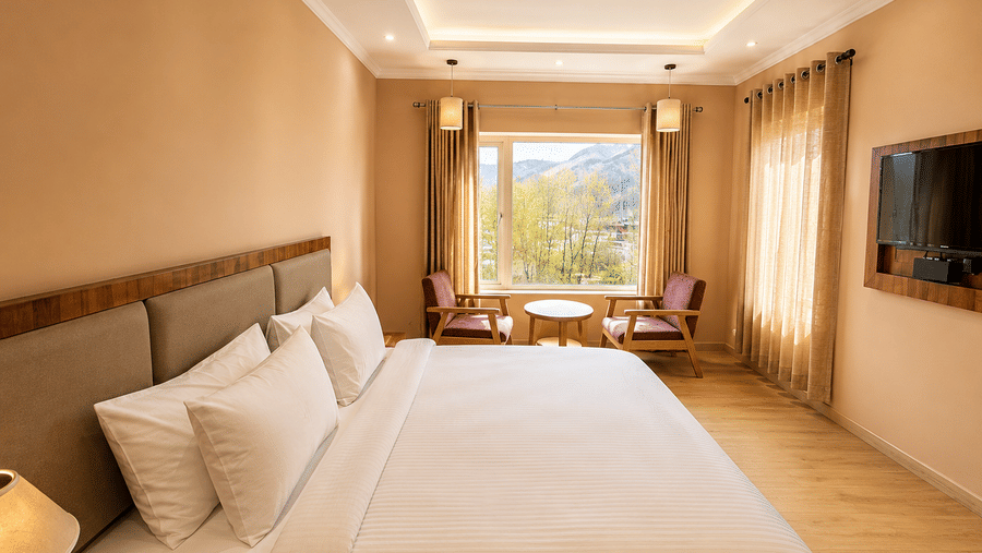Hotel room with a bed dressed in white linen, a seating area with wooden chairs, a wall-mounted television and a large mountain-view window.