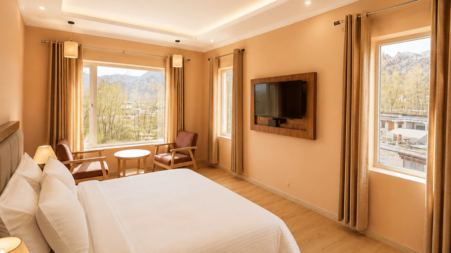 Hotel room with a bed dressed in white linen, a seating area with wooden chairs, a wall-mounted television and long curtains by the window.