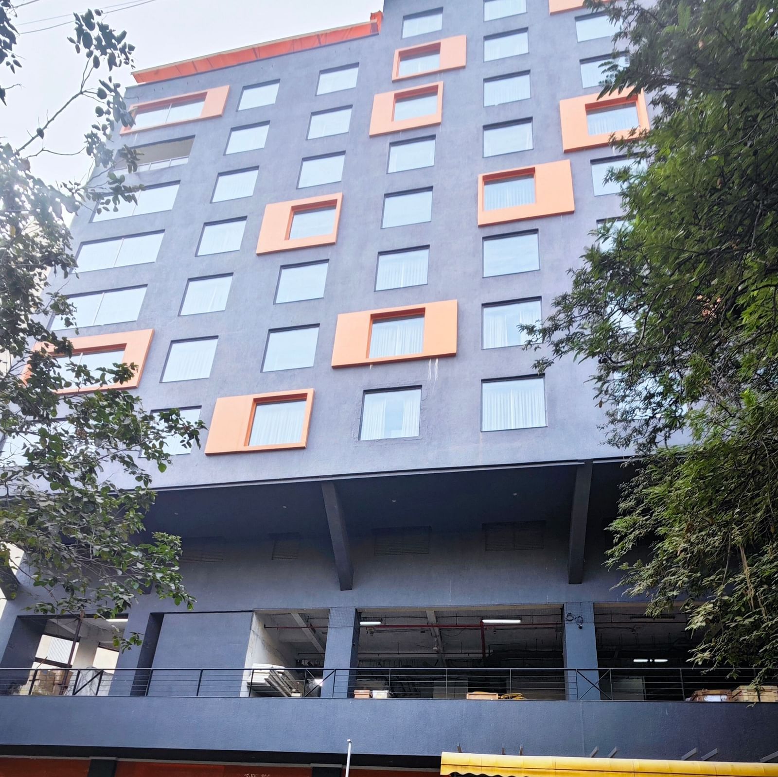 A modern, multi-story hotel building with a dark grey facade and bright orange window accents. The hotel has a covered parking area in front, and there are trees and shrubs surrounding the building.