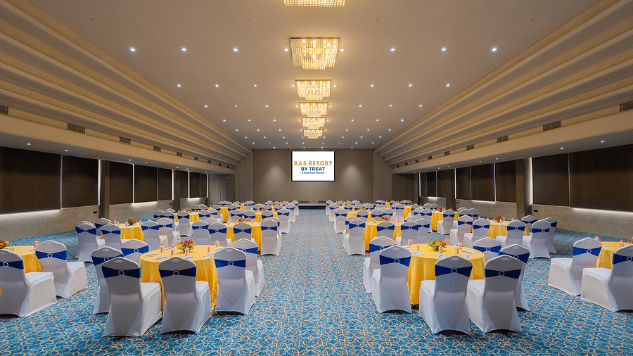 A wide angle view of a spacious banquet hall at Ras Resort by Treat, Silvassa, with round tables and chairs facing towards the stage..