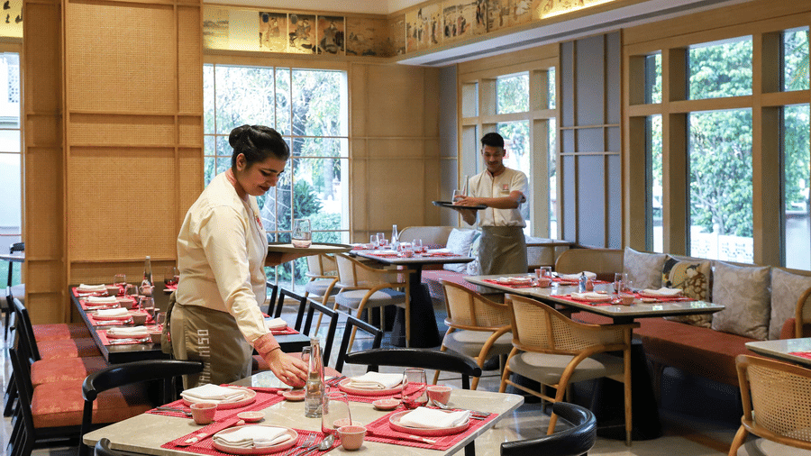 A restaurant interior at Heritage Village Resorts & Spa, Manesar, with staff arranging table settings, placemats, and folded napkins, with another staff member standing near the windows.