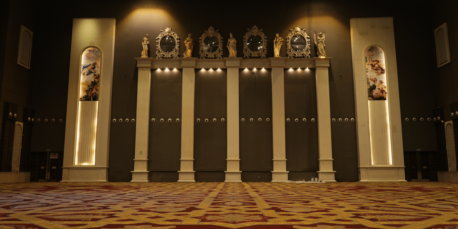 Formal stage backdrop with vertical panels, subtle lighting, and framed portraits or seals displayed high up on the wall at Hotel Hukam's Lalit Mahal.