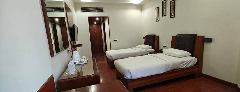 Corner view shot of a bedroom featuring two single beds along with dark wooden flooring with the entrance corridor at the far end of the room - Hotel Gold, Panipat