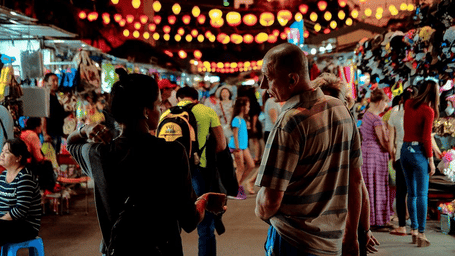 A bustling street market at dusk, adorned with hanging lights, filled with people browsing and shopping.