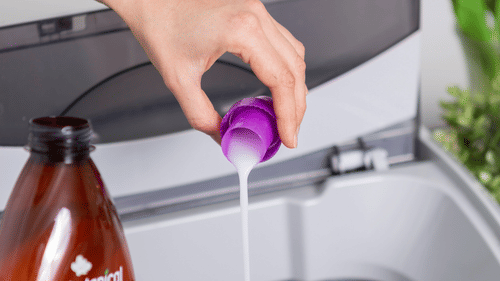 Person pouring plant-based fabric conditioner from a purple cap into a washing machine.