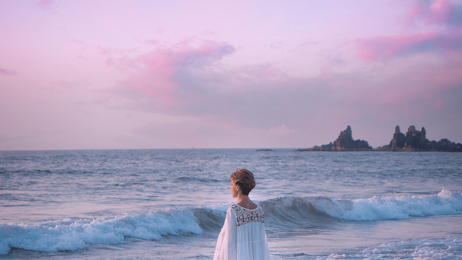 A person in a white dress stands at the edge of the water on a beach, looking out at the ocean under a vibrant pink and blue sunset sky. Distant rock formations are visible in the background.