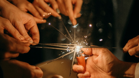 Group of hands holding lit sparklers together at the centre of a table during an evening celebration with bokeh lights in the background.