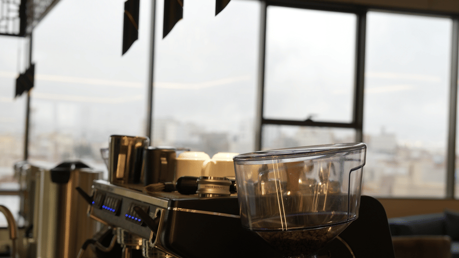 Counter of Grand Iva Cafe at Grand Iva Hotel, Abha, featuring a coffee machine, hanging ambient lighting, and large glass window showcasing the city's views.