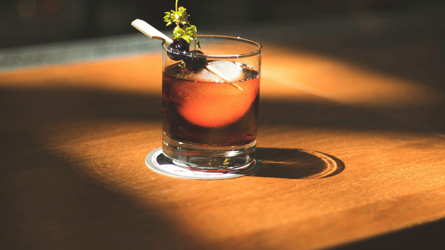 A glass of dark cocktail with ice and garnish, sitting on a wooden bar counter in a sunlit spot.
