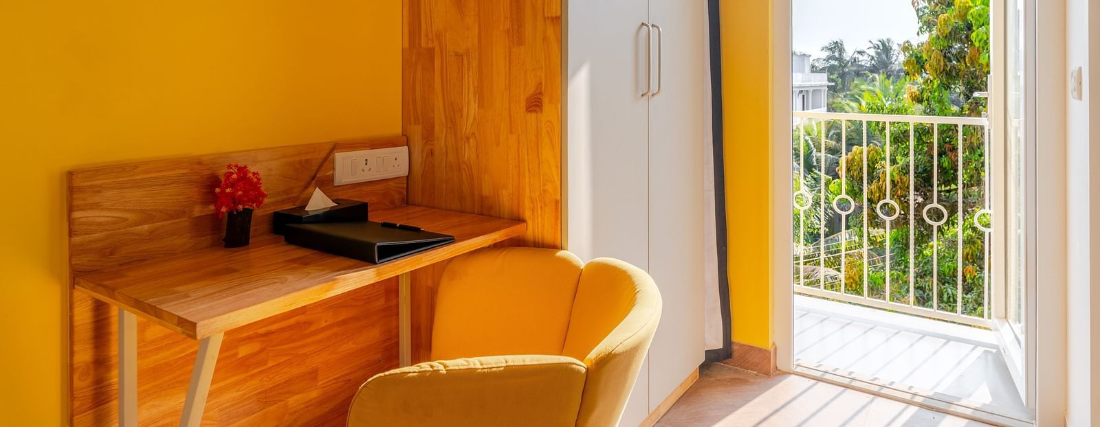 A cozy workspace in a room with yellow walls, featuring a wooden desk and chair set against a window with a view of foliage, creating a serene environment perfect for focus and creativity at Bedzzz Xclusiv Morjim, Goa