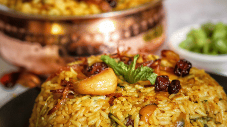 Mound of biryani rice topped with fried cashews, mint and dried berries on a dark plate, with a copper handi of biryani in the background.