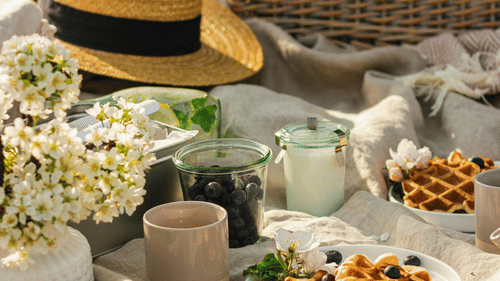 Summer picnic setup on a blanket with a straw hat, waffles, flowers, and a wicker basket.