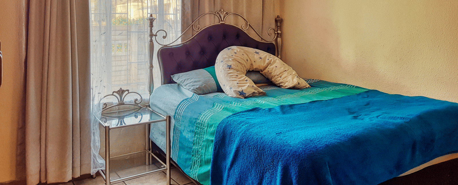 A bedroom featuring a double bed with blue sheets spread neatly over it with beige curtains covering the window behind it.