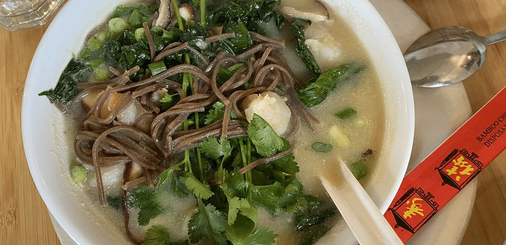 A bowl of soup with noodles, leafy greens, and other ingredients, served with a spoon.