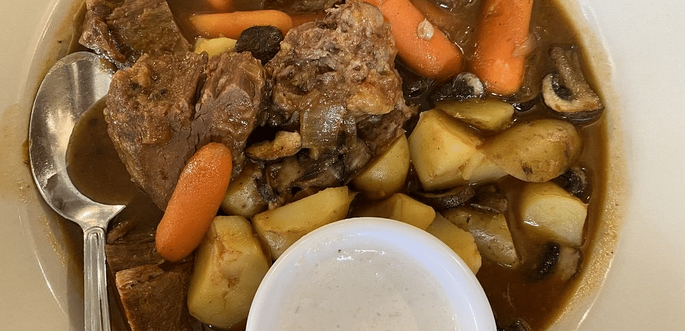 A top view of a bowl of stew with meat, potatoes, and carrots, served with a spoon and a small dish of sauce.