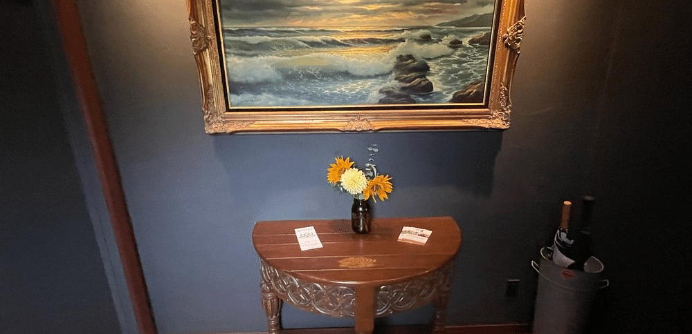 A framed painting on a wall above a small table with a vase and a bottle.