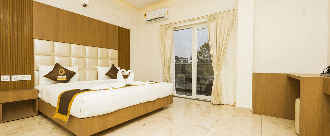 Super Deluxe Room With Balcony featuring a king size bed with a wooden headboard, and decorative pillows, alongside light-coloured walls, tiled flooring.- Sukhi - A Luxury Stay Inn, Chikkamagaluru