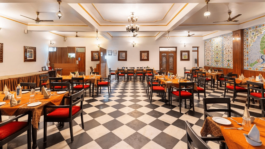 A large indoor restaurant hall featurinf rows of dining tables set on a black and white checkered floor under recessed lights at Amantra Shilpi Resort, Udaipur.