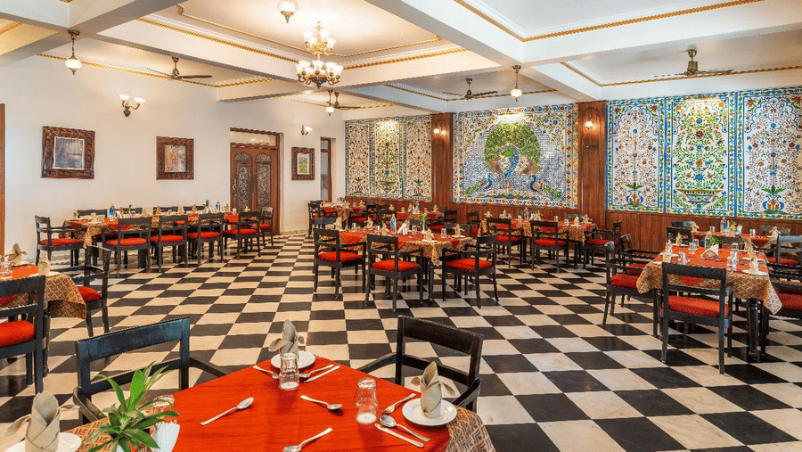 An interior view of a dining hall showing rows of tables with orange cloths arranged on a checkered floor tile pattern at Amantra Shilpi Resort, Udaipur.